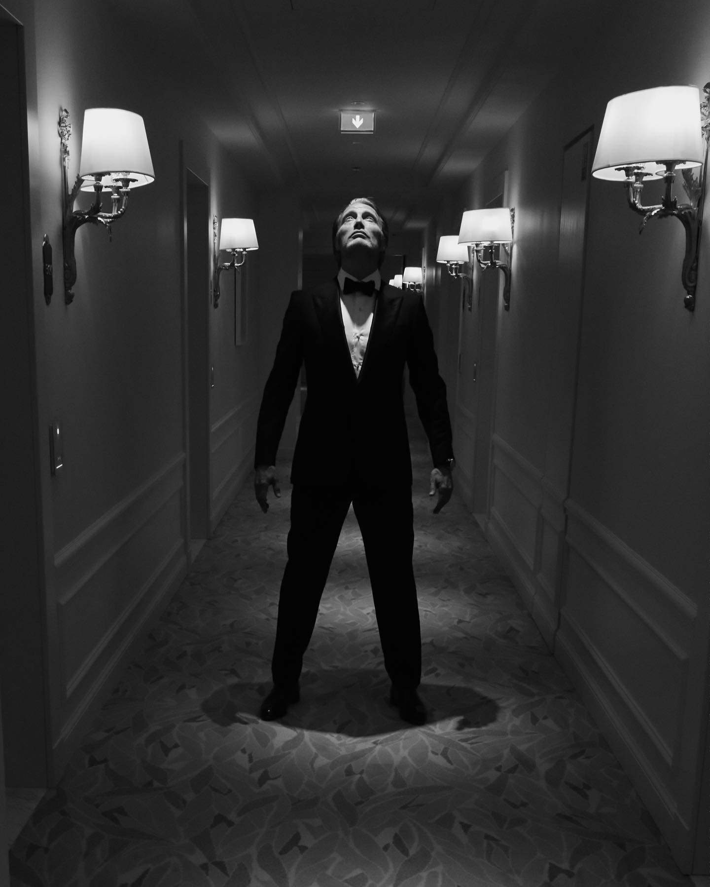 Mads Mikkelsen in a tuxedo standing in a dimly lit hotel corridor, photographed by Greg Williams